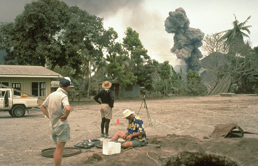 Scientists from the Rabaul Volcano Observatory and the U.S. Geological Survey observe an eruption plume from Tavurvur volcano on 4 October 1994, while conducting deformation measurements on Matupit Island. This location was a tilt site where repeated measurements of uplift or subsidence were made during the course of the eruption. The pit at the lower right was excavated to study ashfall deposits from the eruption. Photo by Elliot Endo, 1994 (U.S. Geological Survey).