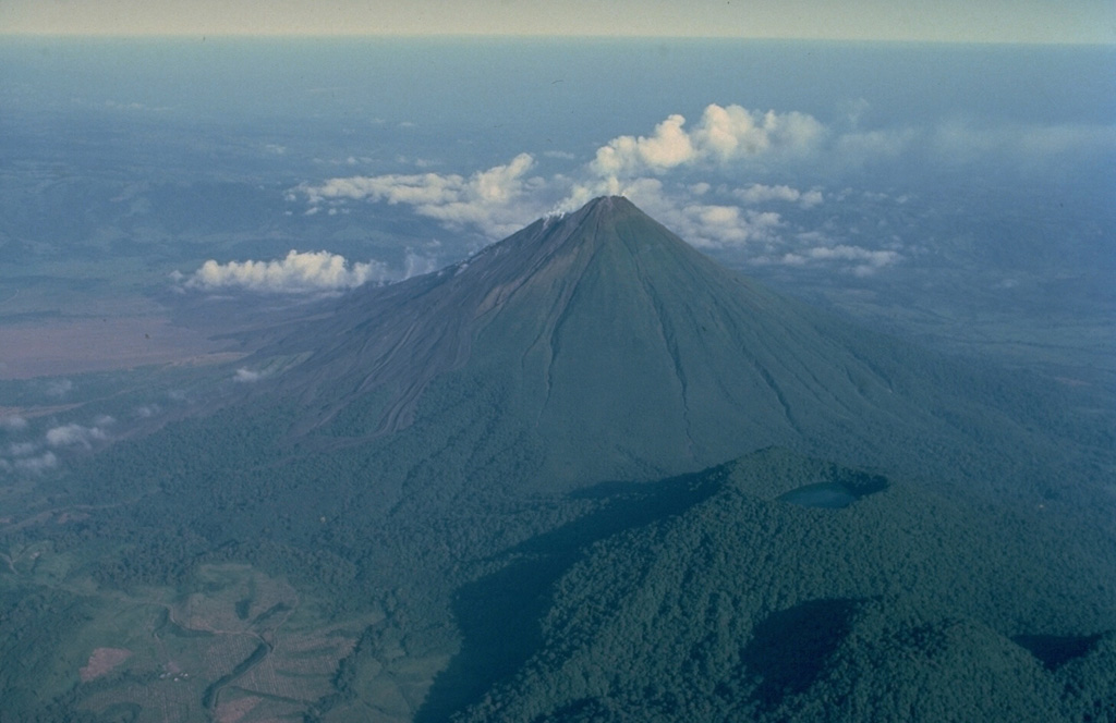 Arenal volcano, seen in this 1983 aerial view from the SE, is a conical stratovolcano that began a long-term eruption in 1968.  Densely forested Cerro Chato (lower right) is an older volcano of the Arenal complex.  Like Arenal, it is armored with lava flows.  The latest eruption of Cerro Chato consisted of phreatomagmatic explosions that formed its summit crater, which is now filled by a lake.  At the extreme lower right is Chatito, a lava dome of the Cerro Chato group. Copyrighted photo by Katia and Maurice Krafft, 1983.