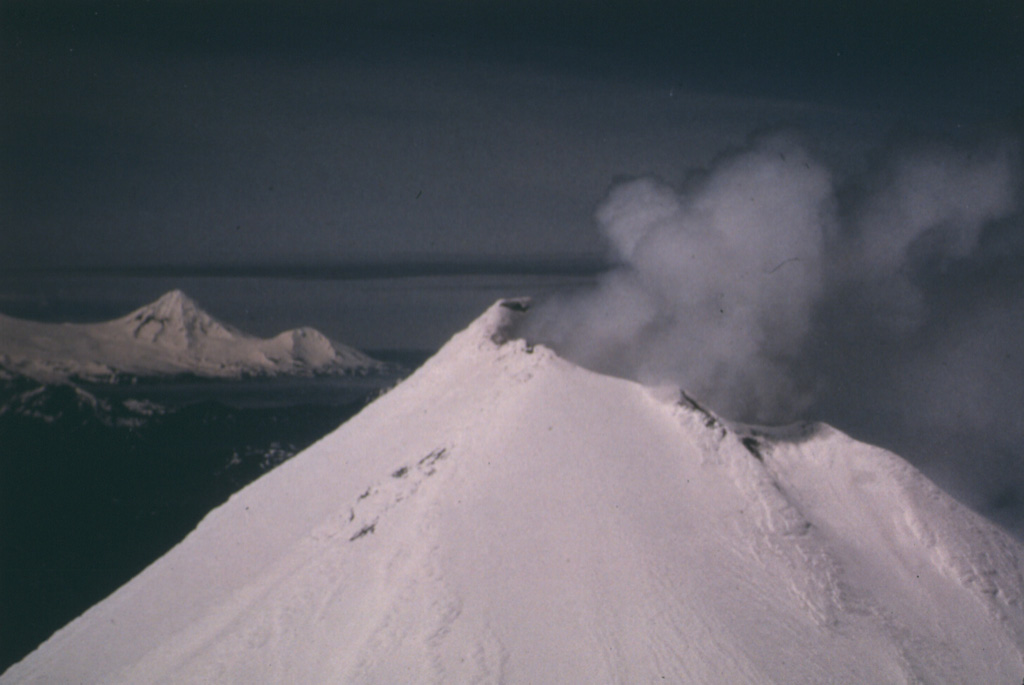 This view of the summit of Shishaldin shows its near- constant crater plume, with its high point on the south rim. Pogromni volcano, part of the Westdahl volcanic complex at the western end of Unimak Island, forms the peak in the distance. Photo Clayton and Marcia Brown, 1987 (courtesy of John Reeder, Alaska Div. Geology & Geophysical Surveys).