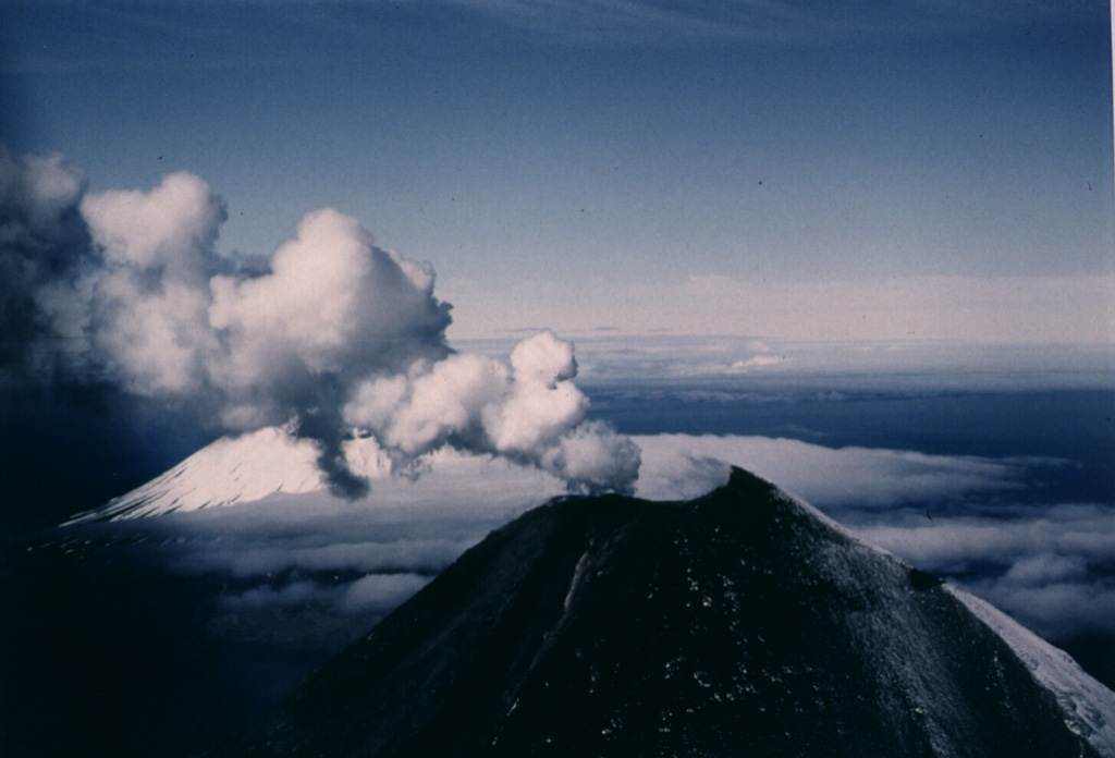 A gas-and-steam plume rises above the summit crater of Cleveland on 23 June 1987. The upper flanks are darkened by ashfall from an eruption that began on 19 June. Lava extrusion began on 23 June and a flow eventually descended 2.5 km down the ESE flank. Lava fountaining was observed on 22 July. A large explosive eruption took place on 28 August, the last day of the eruption. This view from the ENE shows part of Herbert volcano behind the plume to the left.  Photo by Harold Wilson, 1987 (Peninsula Airways), courtesy of John Reeder (Alaska Div. Geology Geophysical Surveys).