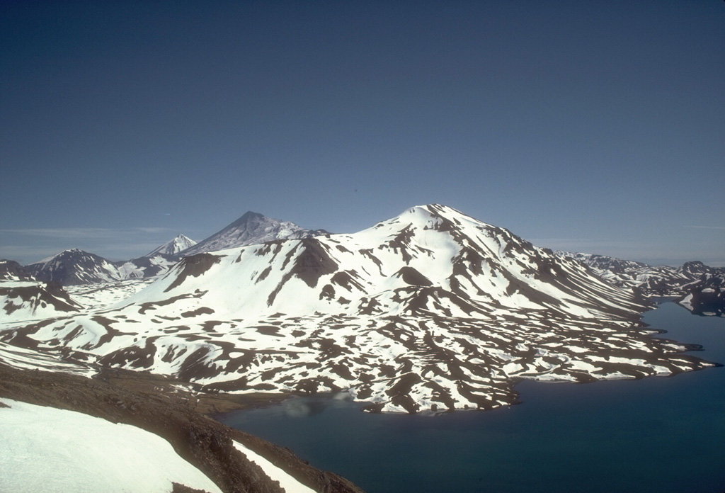 Mount Emmons is along the SW side of the 11 x 18 km Emmons Lake caldera. Youthful lava flows form the irregular shoreline of Emmons Lake in the foreground. The high peaks in the background (left center) are Pavlof just beyond the NE rim of the caldera, and Pavlof Sister. Photo by Tom Miller (Alaska Volcano Observatory, U.S. Geological Survey).
