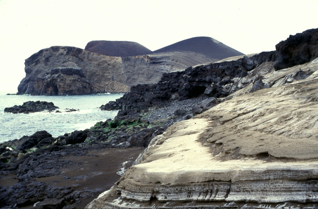The eroded pyroclastic cone at the left, now forming the western tip of Fayal Island, was constructed during the 1957-58 Capelinhos eruption. Submarine eruptions formed an island that was later joined to the mainland, creating a 1-km-wide peninsula. Wave erosion soon truncated the sides of the new cone, forming the steep south-facing cliffs seen here. Beach erosion in the foreground exposes light-colored ashfall deposits from the eruption. Photo by Rick Wunderman, 1997 (Smithsonian Institution).