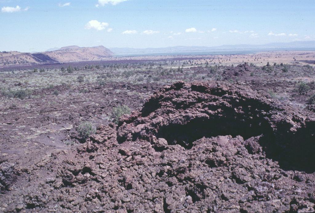 Minor amounts of basaltic lava were erupted about 3000 years ago from spatter cones at Black Crater and Ross Chimneys on the lower northern flank of Medicine Lake volcano.  The spatter cone in the foreground was the vent for the sparsely vegetated small overlapping pahoehoe lava flows in the middle part of the photo.  The dark lava flow in the distance in front of a fault block of older lavas, is the late-Pleistocene Devils Homestead lava flow.   Photo by Lee Siebert, 1988 (Smithsonian Institution).