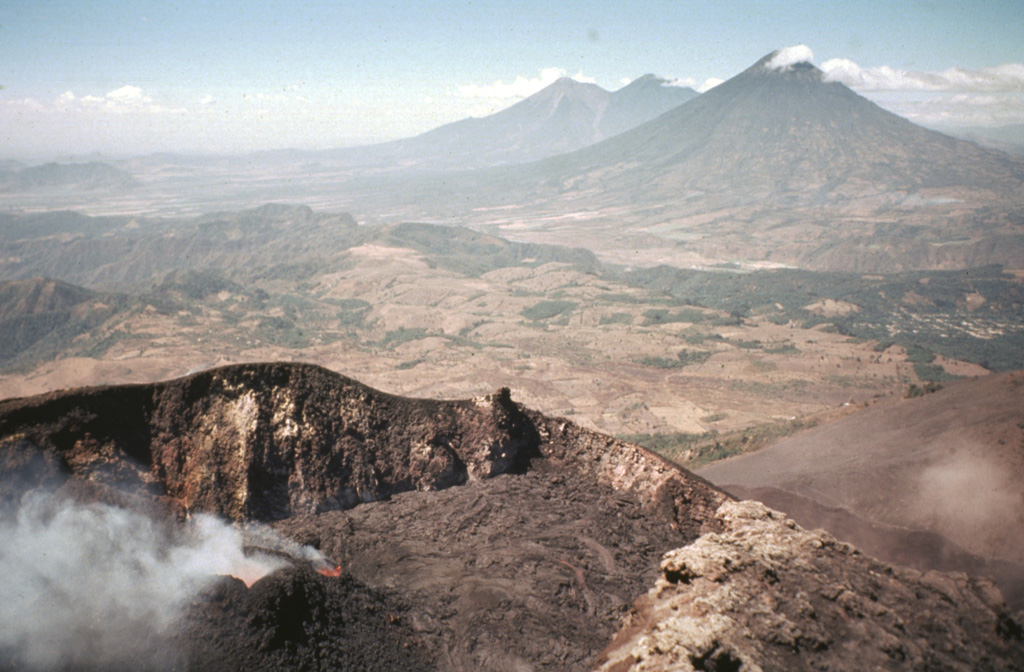 The Pacaya MacKenney crater contains spatter cones that have produced lava flows, and gas plumes like the one seen here. This photo taken in May 1981 shows the broad several-hundred-meter wide crater rim. From 9 May to 2 June lava flows from the spatter cones covered the crater floor and flowed through notch in the NW crater rim. In the background are Fuego and Acatenango (right-center) and Volcán de Agua (right).  Photo by Bill Rose, 1981 (Michigan Technological University).