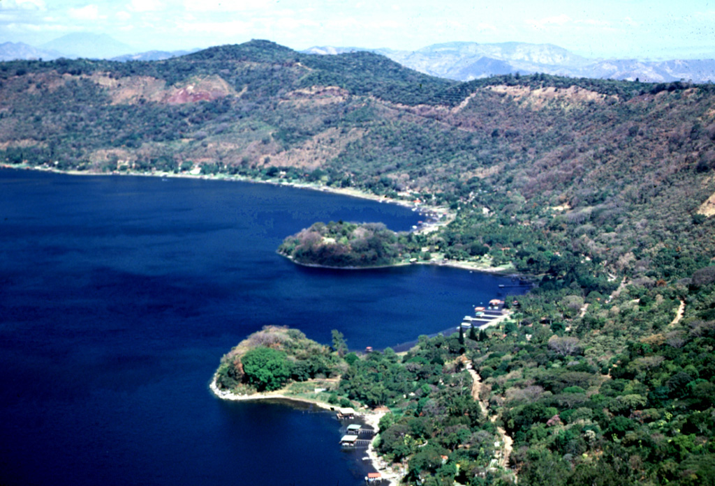 The Los Anteojos lava domes form small peninsulas on the eastern shore of Lago de Coatepeque. Four other domes are on the opposite side of the caldera in a trend that passes through Izalco volcano to the SW. The Anteojos domes are mostly submerged, rising only about 20-25 m above the lake surface in this 2002 photo. A chain of scoria cones was erupted along caldera ring faults; two of these are seen beyond the two domes. Photo by Lee Siebert, 2002 (Smithsonian Institution).