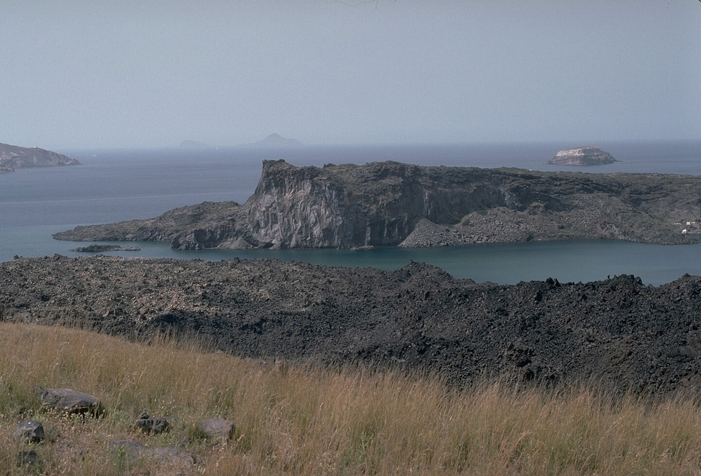 Palaea Kameni (Thia) Island (center) was formed by the extrusion of lava flows during a 46-47 CE eruption. This was the second documented eruption producing a new island in the caldera bay. The black lava flows in the foreground on Nea Kameni island were emplaced during an 1866-1870 eruption. The small island in the right distance to the SW, capped by light-colored rocks of the 3,600-year-old Minoan eruption, is part of the caldera wall that extends beneath the sea surface to the Akrotíri Peninsula on the extreme left. Photo by Lee Siebert, 1994 (Smithsonian Institution).