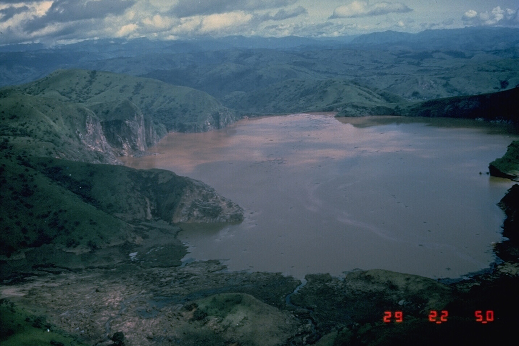 Lake Nyos is the most renowned of the numerous maars and basaltic cinder cones associated with the deeply dissected Mount Oku massif. The 1.2 x 1.9 km wide lake, seen here from the south, was the site of a gas-release event on 21 August 1986 that caused at least 1,700 fatalities. Wave damage stripped the peninsula at the left of vegetation. The emission of around 1 km3 of magmatic carbon dioxide has been attributed to either non-volcanic overturn of stratified lake waters, to phreatic explosions, or to injection of hot gas into the lake. Photo by Jack Lockwood, 1986 (U.S. Geological Survey).