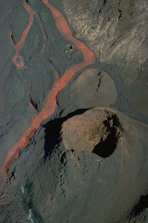 An incandescent lava channel diverts around a lava cone on Piton de la Fournaise volcano in September 1985.  A long-term eruption beginning in June 1985 produced more than 0.1 cu km of lava flows from fissures in the summit crater, the flanks of the central lava shield, and the outer flanks of the caldera. Copyrighted photo by Roland Benard, 1985 (courtesy of Katia and Maurice Krafft).