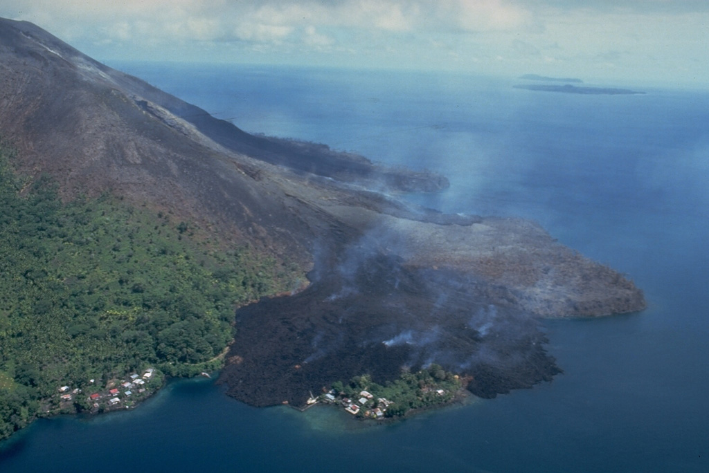 This May 19, 1988, view from the NE shows a still-steaming black lava flow entering the sea along the north coast, partially overrunning the village of Batu Angus.  The lava flow originated from a vent at 200 m on the north flank on May 9, and reached the sea the same day.  A second steaming lava flow, the Pasir Besar flow, which originated from a vent at 300-350 m elevation, can be seen reaching the NW coast at the center of the photo.  The portion of Batu Angus village surrounded by the lava flow sits on an earlier lava flow erupted in 1901. Photo by Tom Casadevall, 1988 (U.S. Geological Survey).