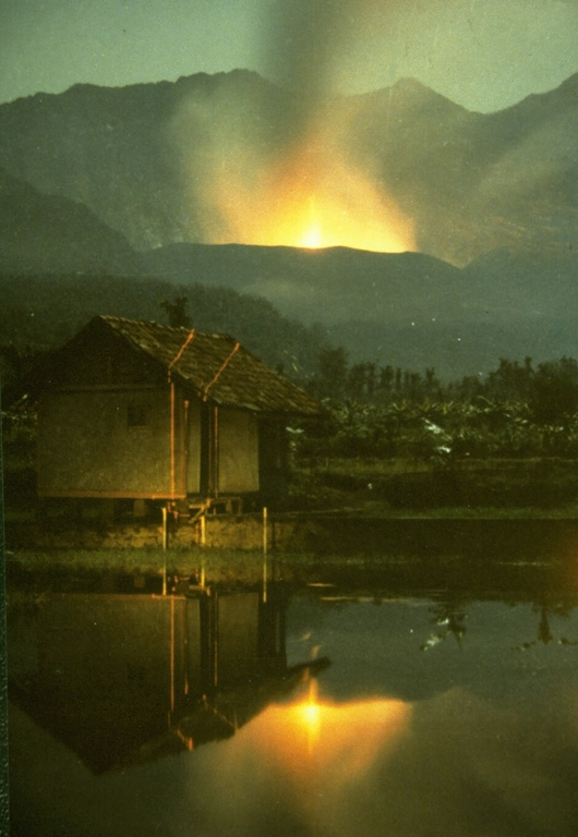 Strombolian eruptions from the crater of Galunggung volcano on 23 December 1982 are reflected in a pond at Kubanghurang village, SE of the volcano. The vertical orange lines crossing the house at the left are the traces of an Electronic Distance Measurement (EDM) laser used by Volcanological Survey of Indonesia scientists to monitor deformation of the volcano. Photo by Ruska Hadian, 1982 (Volcanological Survey of Indonesia).