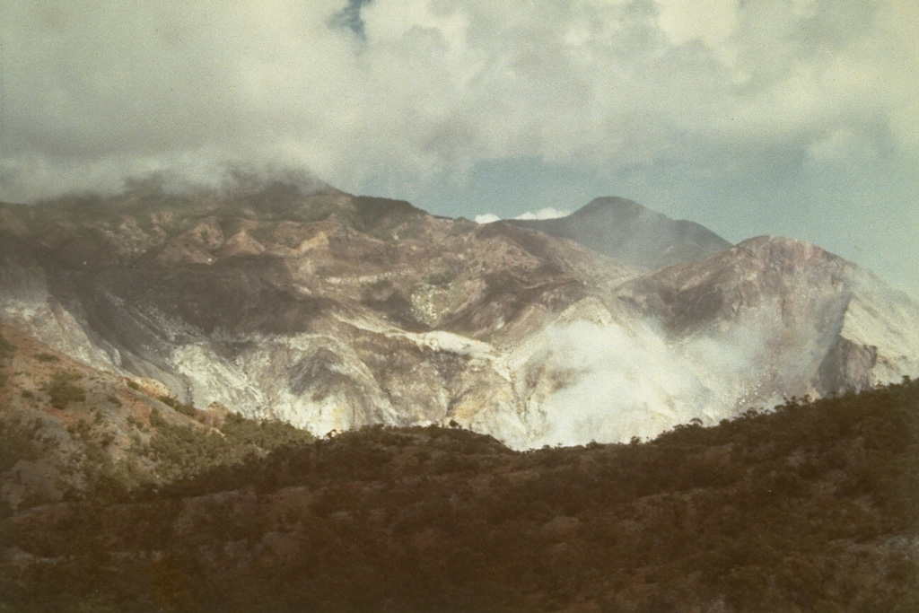 Steam clouds rise above a fumarole field on the flank of Gunung Sirung, south of Airmama village.  The crater walls contain extensive areas of light-colored hydrothermally altered rock.  Photo by L.D. Reksowirogo, 1972 (Volcanological Survey of Indonesia).