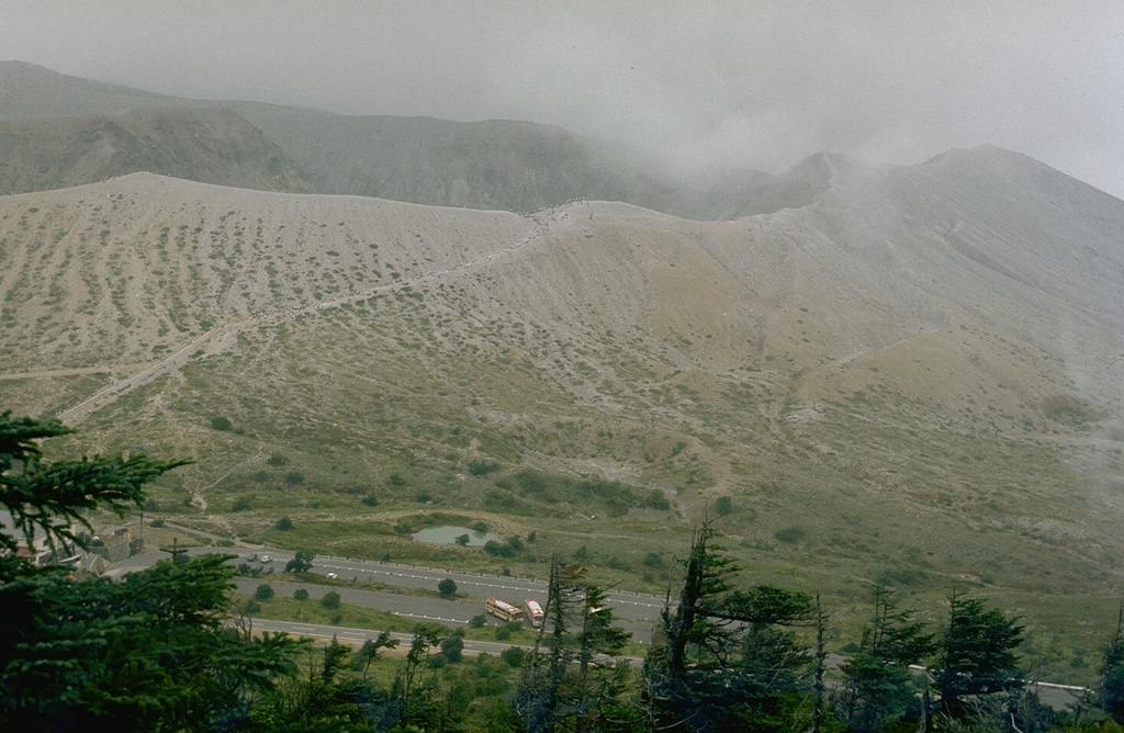 The crater rim of Kusatsu-Shiranesan is seen here from the top of a flank cone to its S. The slopes of the cone are kept largely unvegetated by frequent phreatic explosions from three overlapping craters. Eruptions have been recorded since the early 19th century. Photo by Lee Siebert, 1977 (Smithsonian Institution).