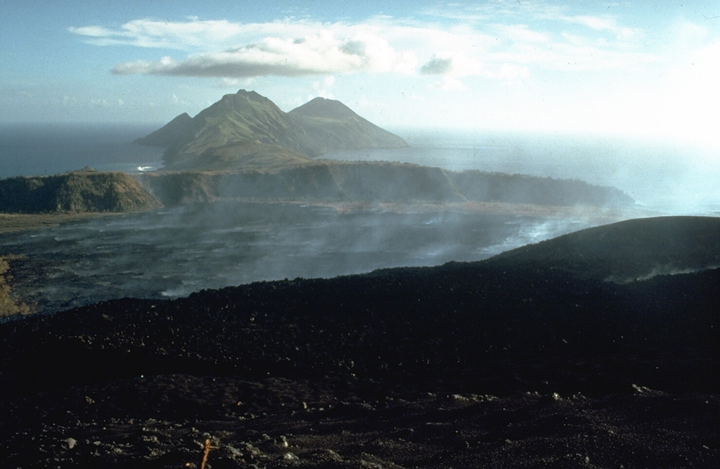 Gases rise from a lava flow that is advancing below the S flank of Pagan in 1981. The flow erupted during 15-26 May 1981 from a vent on the S crater rim and spread out over the broad caldera floor, eventually reaching the southern caldera wall. The foreground area on the flanks of Pagan consists of pyroclastic surge deposits from the 1981 eruption. This 21 May 1981 photo looks to the SW with South Pagan volcano forming the conical peak at the right. Photo by Norm Banks, 1981 (U.S. Geological Survey).
