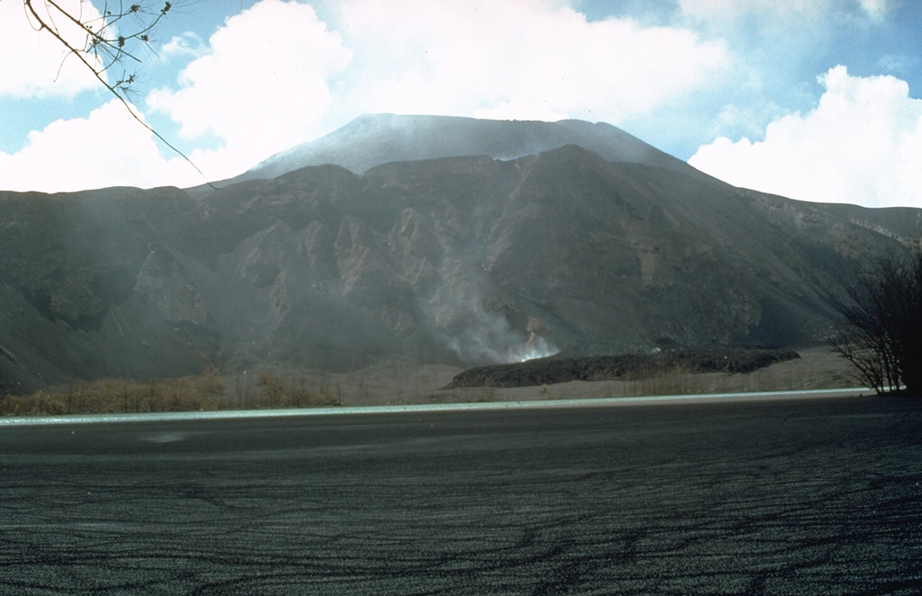 A black lava flow advances across the floor of a maar on the W flank of North Pagan volcano on 23 May 1981. The flow originated from the summit crater of North Pagan (background) on 15 May. Most of the surface of a lagoon within the maar in the foreground is covered by black floating material from the explosive eruption on the 15th. Photo by Norm Banks, 1981 (U.S. Geological Survey).