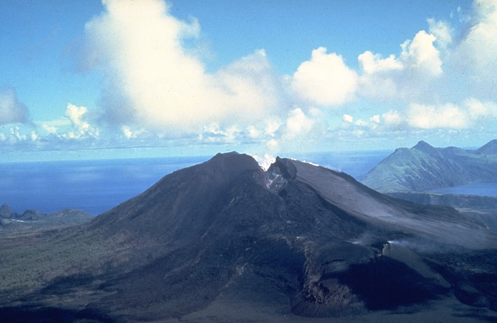 A fissure that formed during an eruption of North Pagan volcano in the Mariana Islands in 1981 cuts across the summit of the volcano. Three principal vents were active along the fissure. A scoria cone (foreground) was constructed at the northernmost vent, and vents on the north and south rims of the summit crater fed lava flows that traveled down the flanks. Blocks as large as 1 m in diameter were ejected to distances of 2 km from the vent. This 16 June 1981 photo shows South Pagan volcano at the upper right. Photo by U.S. Navy, 1981.