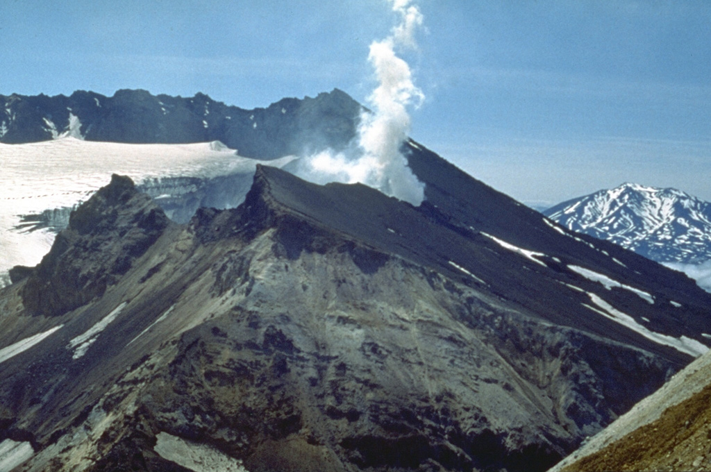 The glacier-filled southern summit crater of Mutnovsky is seen here from the NE with Asacha in the distance. This crater is within a larger 1.5 x 2.1 km wide crater with walls 50-250 m high. Four smaller craters are on the northern rims of the summit crater complex. A steam plume rises from the historically active crater on the western rim of northern Mutnovsky crater. Photo by Oleg Volynets (Institute of Volcanology, Petropavlovsk).