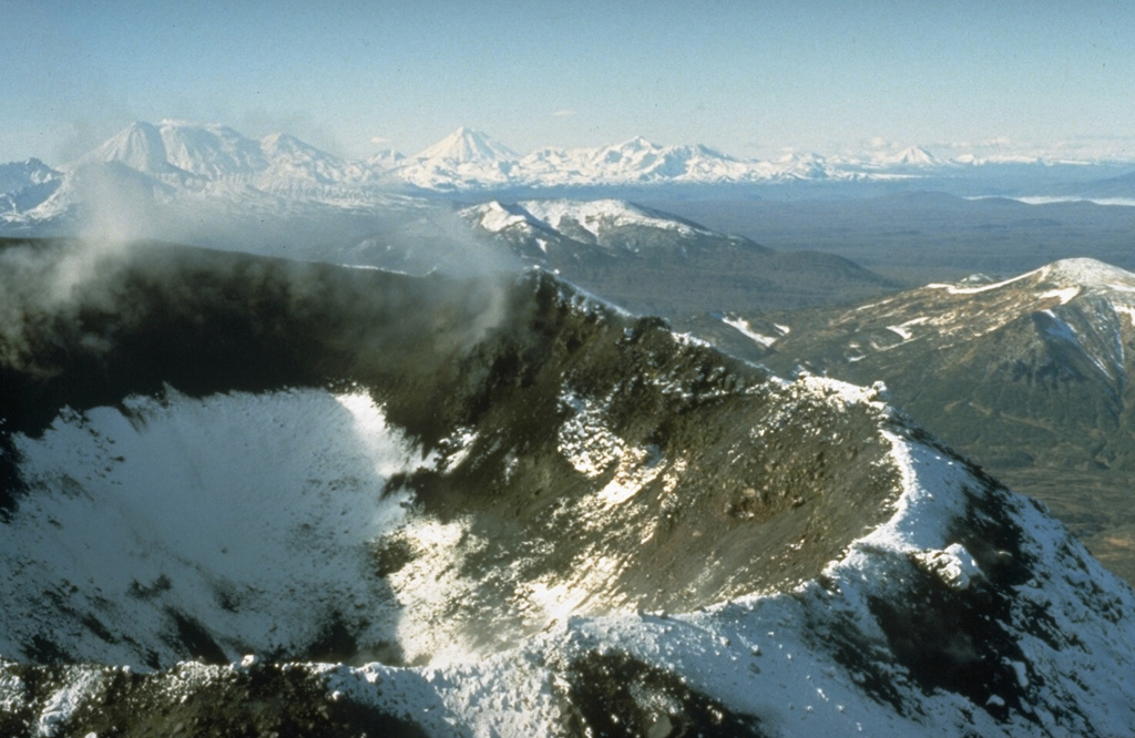 The 200-250 m wide Karymsky summit crater formed following a long-term eruption during 1970-82. This early 1990's photo shows the crater from the NE. Several other prominent central Kamchatka volcanoes are in the background. Zhupanovsky, to the far left, is composed of four overlapping edifices constructed along an WNW-ESE line. Koryaksky is to the left-center, and the broad Dzenzursky massif to its right. Photo by Dan Miller (U.S. Geological Survey).