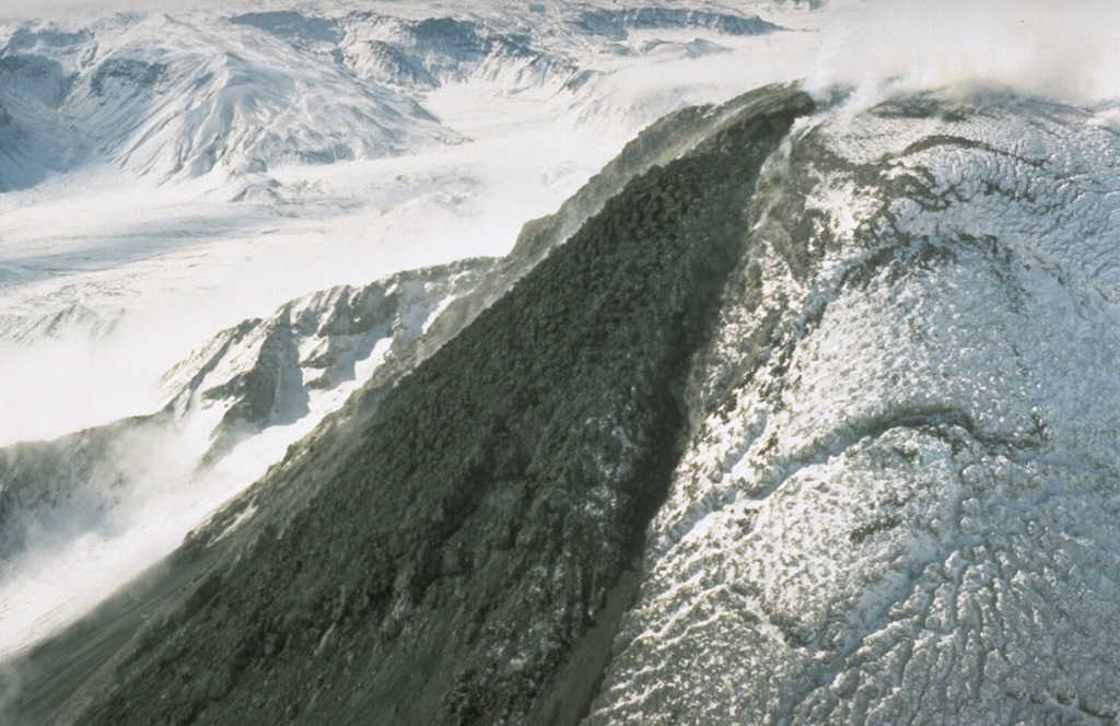 A black lava flow descends from the snow-mantled summit lava dome of Bezymianny volcano in Kamchatka in September 1990. The lava dome formed during the past forty years both by expansion when new magma was intruded into the dome and by the extrusion of lava flows down its flanks. The dome has grown within a large crater, whose southern rim is visible behind the lava flow. Photo by Dan Miller, 1990 (U.S. Geological Survey).