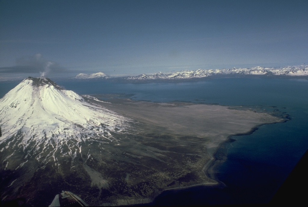 A small plume rises from the summit of glaciated Augustine in this July 1988 view from the east. The gray, unvegetated areas on the north flank are pyroclastic flow deposits from the 1986 and earlier historical eruptions. The pyroclastic flows reached the sea to the east and west. The Chigmit Mountains rise across Kamishak Bay in the distance. Photo by Bill Rose, 1988 (Michigan Technological University).