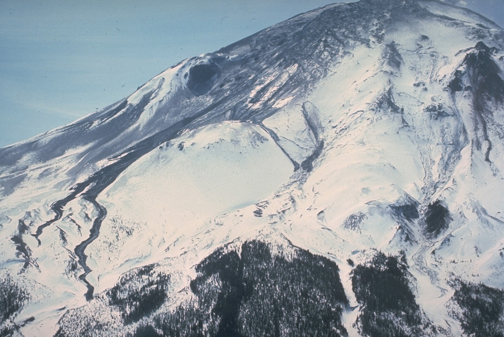 On 11 April 1980, two weeks after the 27 March beginning of the eruption of Mount St. Helens, an area of fractured glacial ice is visible at the top of the volcano. Thin black streaks of volcanic mudflows are visible at the center and lower left that diverge around the undisturbed snowpack on Sugar Bowl lava dome. Photo by William Melson, 1980 (Smithsonian Institution).