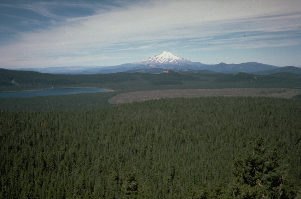 Medicine Lake volcano is seen here looking over the summit crater from the east with Mount Shasta in the background. The barren Medicine Glass Flow on the right was erupted about 5,200 years ago. During the Holocene, obsidian flows were erupted from summit and flank vents, and voluminous basaltic lava flows originated from vents on the north and south flanks. Photo by Dan Dzurisin, 1985 (U.S. Geological Survey).