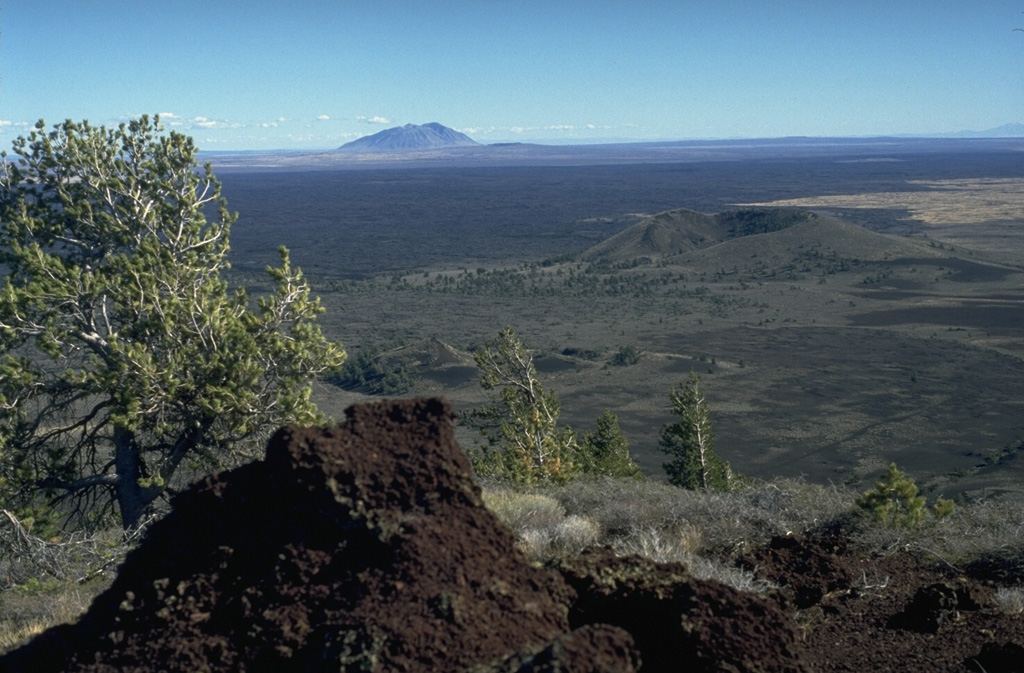 The vast extent of the Blue Dragon lava flow, forming the flat area in the middle of the photo, can be appreciated in this view from Big Cinder Butte with Big Southern Butte in the background to the east.  The Blue Dragon flow, the largest volume lava flow at Craters of the Moon, covers 280 km2 with 3.4 cu km of lava.  The flow was erupted about 2075 years ago and covers broad areas as far as 25 km to the east and 15 km to the SW of its vent area. Photo by Lee Siebert, 1994 (Smithsonian Institution).