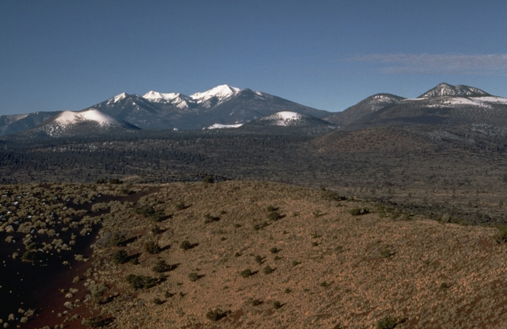 The snow-drapped symmetrical cinder cone at the left side of the photo is Sunset Crater, Arizona's most recently active volcano.  The broad snow-capped mountain in the background to the west, behind and to the right of Sunset Crater, is San Francisco Mountain.  It is the centerpiece of the San Francisco Mountain volcanic field, which covers 5000 km2 of northern Arizona.  The massive eroded Pleistocene stratovolcano is Arizona's highest peak.  The peak on the right horizon is O'Leary Peak, a Pleistocene rhyodacitic lava dome complex.   Photo by Ed Wolfe, 1970 (U.S. Geological Survey).