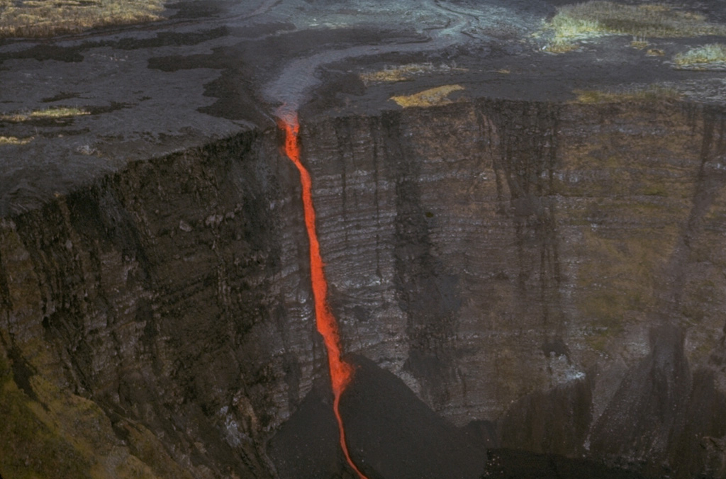 A stream of lava from a vent at Mauna Ulu (above the top of the photo) cascades into Makaopuhi crater during an early stage of the 1969-74 Mauna Ulu eruption. A thick stack of older lava flows that were erupted along Kilauea's East Rift Zone is exposed in the Makaopuhi crater wall. Photo by Jim Moore (U.S. Geological Survey).