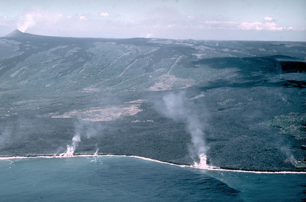 Laze (a plume of hydrochloric acid, steam, and fine glass particles) rises along the Puna coast where lava flows enter the sea in this 28 December 1987 photo. The flows originated from a fissure along Kilauea's East Rift Zone from the Pu‘u ‘Ō‘ō scoria cone (upper left) to the Kūpaianaha lava lake (small gas plume in the center of the horizon). Lava flows traveled 10 km to the coast and inundated several subdivisions and villages. Photo by J.D. Griggs, 1987 (U.S. Geological Survey).