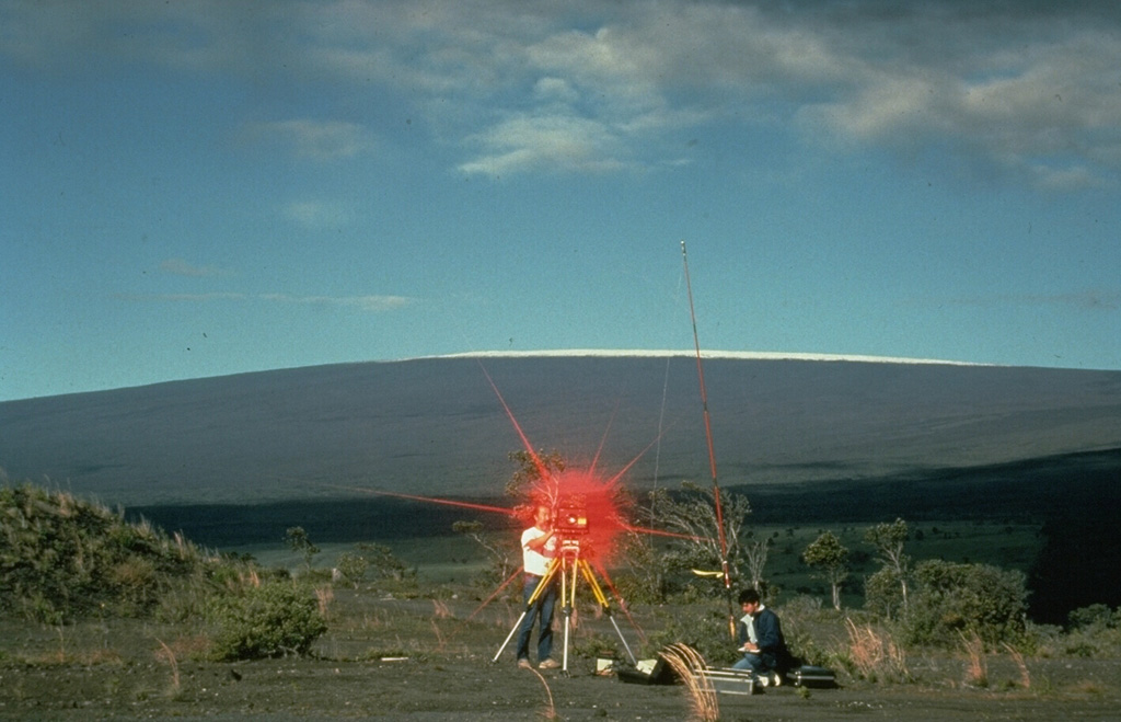 Hawaiian Volcano Observatory scientists conduct an electronic-distance measurement (EDM) survey on the rim of Kilauea caldera in 1988, with snow-capped Mauna Loa in the background. The procedure uses a laser beam, which is reflected back to the EDM instrument from a distant cluster of reflectors. A precise determination of the distance between the two points is made by a small computer in the EDM instrument. These measurements allow scientists to detect inflation or deflation of the volcano due to changes in the magmatic or hydrothermal systems. Photo by J.D. Griggs, 1988 (U.S. Geological Survey).