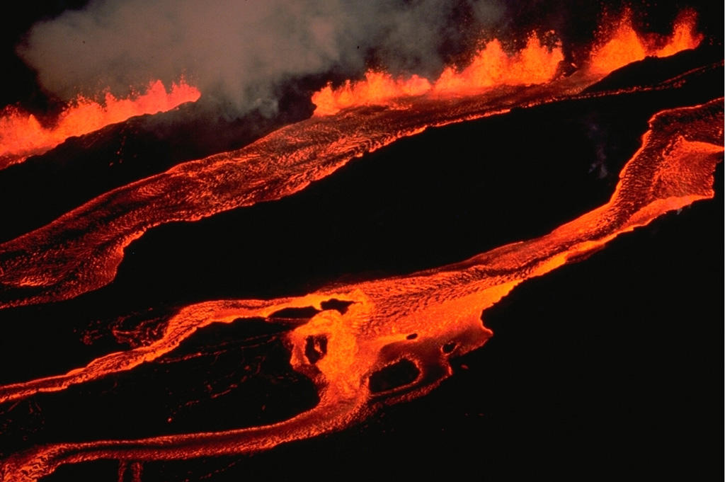 The en-echelon character of the 1984 eruptive fissure at Mauna Loa volcano can be appreciated in this March 25 view from the north along the NE rift zone.  The lava fountain at the upper left feeds a lava flow that travels downslope out of view to the left.  In the center, a lava flow can be seen that originated from the adjacent offset fissure segment immediately up slope, while the flow to the right originates from another fissure segment farther up slope beyond the photo margin. Copyrighted photo by Katia and Maurice Krafft, 1984.