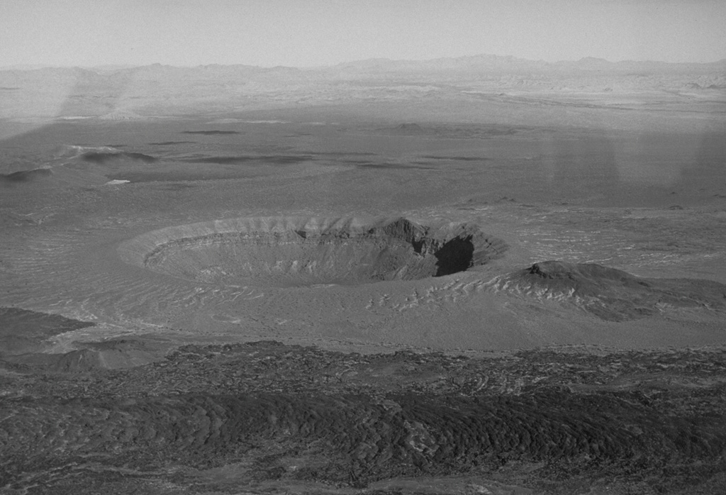 Cráter Elegante is one of the largest maars of the Pinacate volcanic field, seen here from the WSW. The uneven area on the southern crater rim (to the right) is a scoria cone that was dissected during formation of the maar. Another darker scoria cone to the right is surrounded by lighter-colored pyroclastic surge deposits from the maar-forming eruptions. A third scoria cone (lower left) opens toward the rim of Cráter Elegante and is partially surrounded by the younger dark-colored lava flow in the foreground. Photo by David Roddy, 1965 (U.S. Geological Survey).