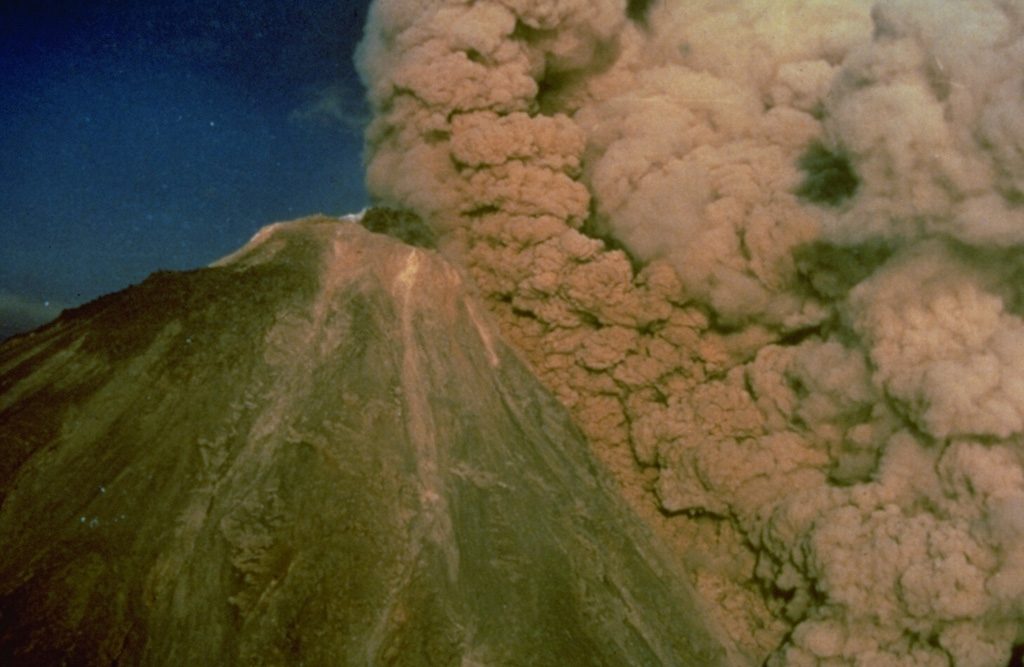 An ash plume rises above a pyroclastic flow traveling down the SW flank of Colima on 16 April 1991, colored orange by the late-afternoon sun. The pyroclastic flow was produced by collapse of unstable parts of the summit lava dome. The black mass at the summit is a lava dome that began growing on 1 March. Later in the eruption, which ended in October, a lava flow traveled down the SW flank to 2,600 m elevation from the roughly 3,850-m-high summit. Photo by Alfredo Ramirez (pilot Ernesto Gómez Hofman), 1991 (courtesy Melchor Urzua, Protección Civil de Colima).
