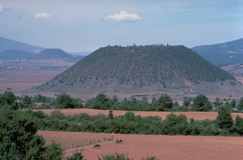 The flat-topped Hoya el Huanillo cinder cone, located about 35 km NE of Parícutin, erupted during the early Holocene about 9300 years ago.  It is seen here from the NW with the town of Cherán in the left distance near the Cerro Cucundicata cinder cone.  In contrast to many other Michoacán cinder cones, only a small ash-covered lava flow was emitted from Hoya el Huanillo. Copyrighted photo by Katia and Maurice Krafft, 1981.