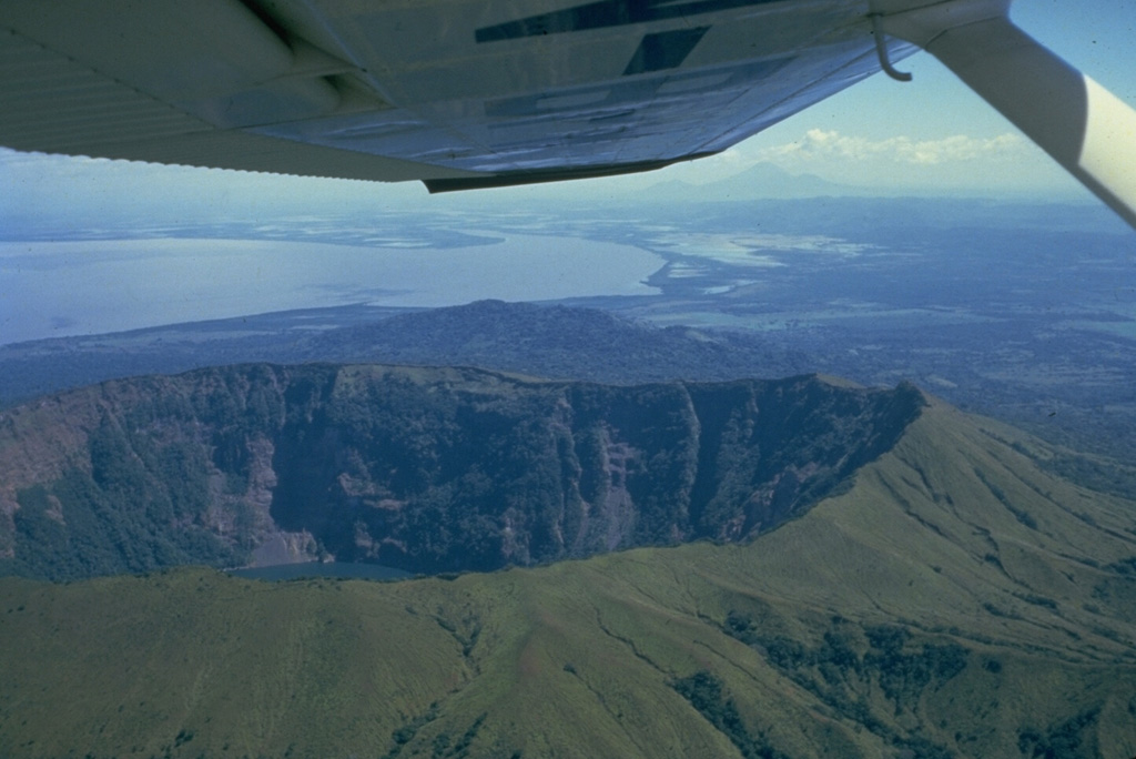 The summit of Cosigüina volcano, seen here from the west with the Gulf of Fonseca in the background and the border with Honduras at the upper left, contains a 2 x 2.4 km wide caldera with steep-sided, 500-m-high walls.  The upper flanks of the volcano are only partially forested after a devastating eruption in 1835 produced pyroclastic flows and surges that reached the coast and fountain-fed lava flows that blanketed the upper eastern and western flanks.  The peak in the distance below the right side of the wing is San Cristóbal volcano. Photo by Jaime Incer, 1981.