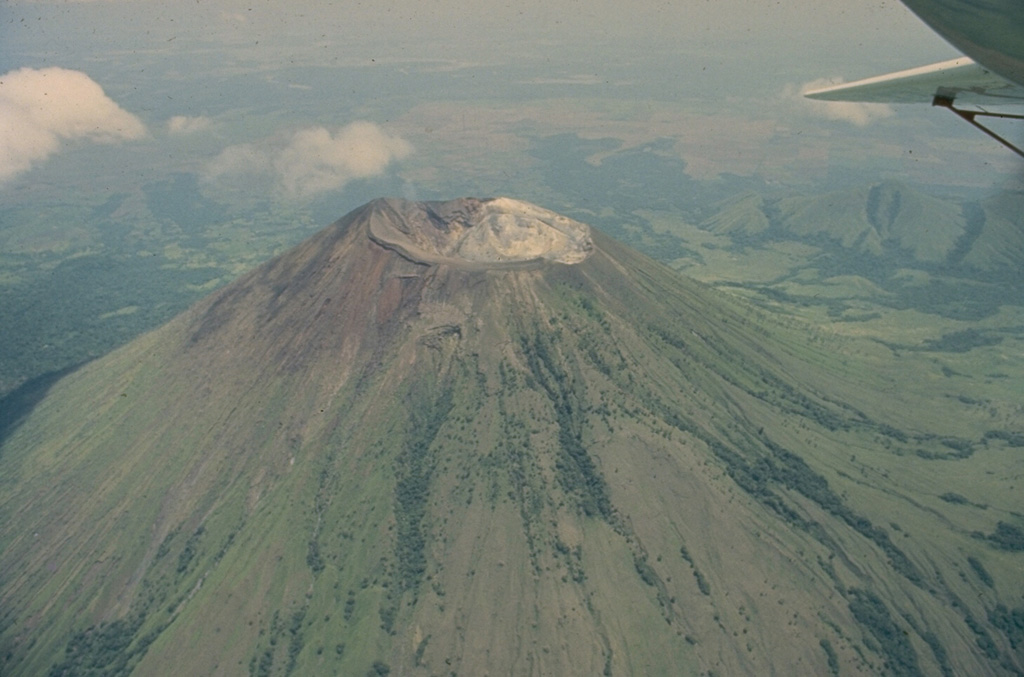 San Cristóbal, seen in this 1975 aerial view from the south, is the highest volcano in the Marrabios Range of western Nicaragua.  The symmetrical, 1745-m-high stratovolcano is the youngest of five principal volcanoes forming the San Cristóbal complex.  Young lava flows are located on its SW and northern flanks.  Explosive eruptions have occurred since the beginning of the Spanish era.  Cerro Moyotepe, a small composite volcano of the San Cristóbal complex that is cut by a series of N-S faults, appears at the right, below the plane wing. Photo by Jaime Incer, 1975.