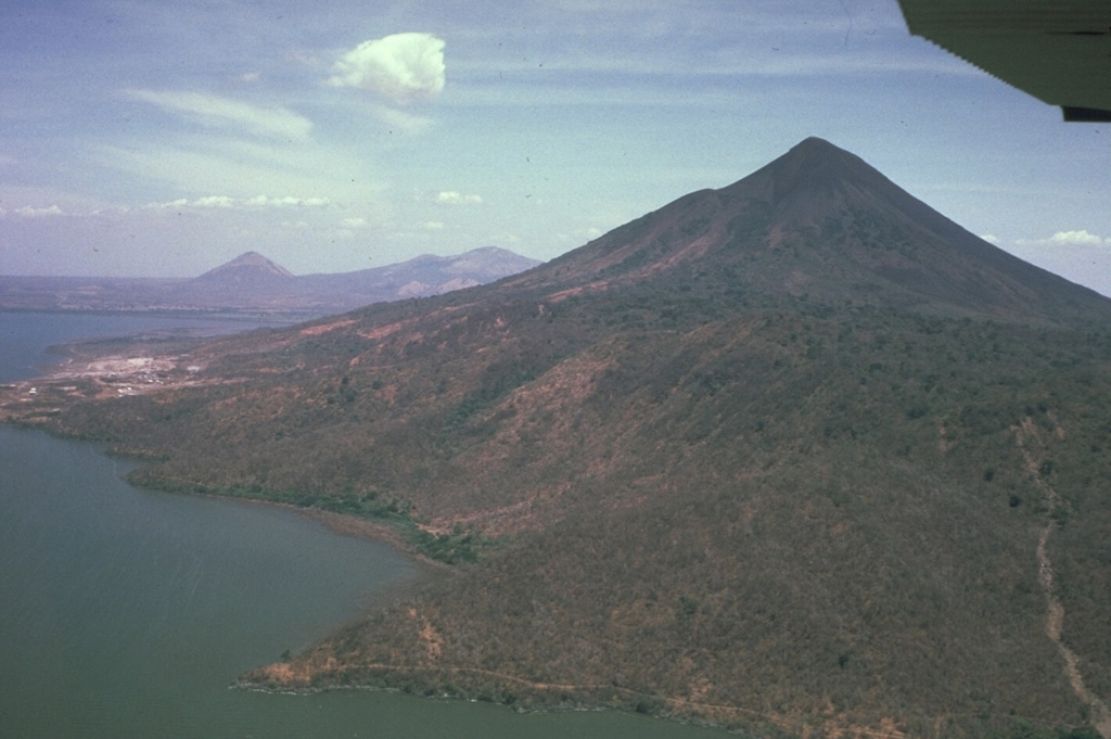 Momotombo volcano, seen here from the east, is a prominent youthful stratovolcano constructed along the shores of Lake Managua.  The peninsula in the foreground is Punta del Diablo, and the light-colored area at the left is the site of the Momotombo geothermal plant.  The young volcano is only 4500 years old and has a long record of strombolian eruptions, with occasional larger explosive activity.  In the distance are the conical peak of Cerro Asososca and the broad summit of Las Pilas volcano. Photo by Jaime Incer, 1983.