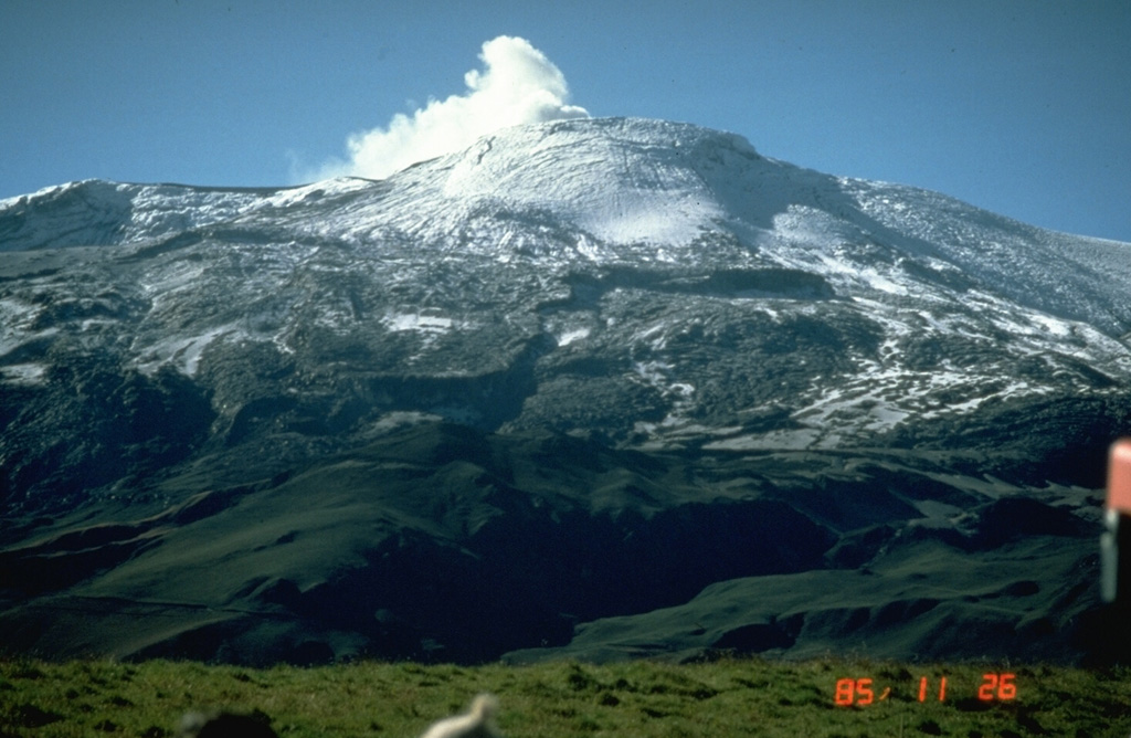 Two weeks after the 13 November 1985 eruption, a plume rises from Las Arenas, the summit crater of Nevado del Ruiz. This view from the NNW shows pyroclastic surge and pyroclastic flow deposits mantling the summit icecap. Grooves descending the upper glacier surface were scoured by pyroclastic surges, which reached 5.5 km NW and NE of the summit. Melting of the summit icecap during the eruption led to devastating lahars that traveled as far as 100 km from the volcano and caused 24,000 fatalities. Photo by Norm Banks, 1985 (U.S. Geological Survey).
