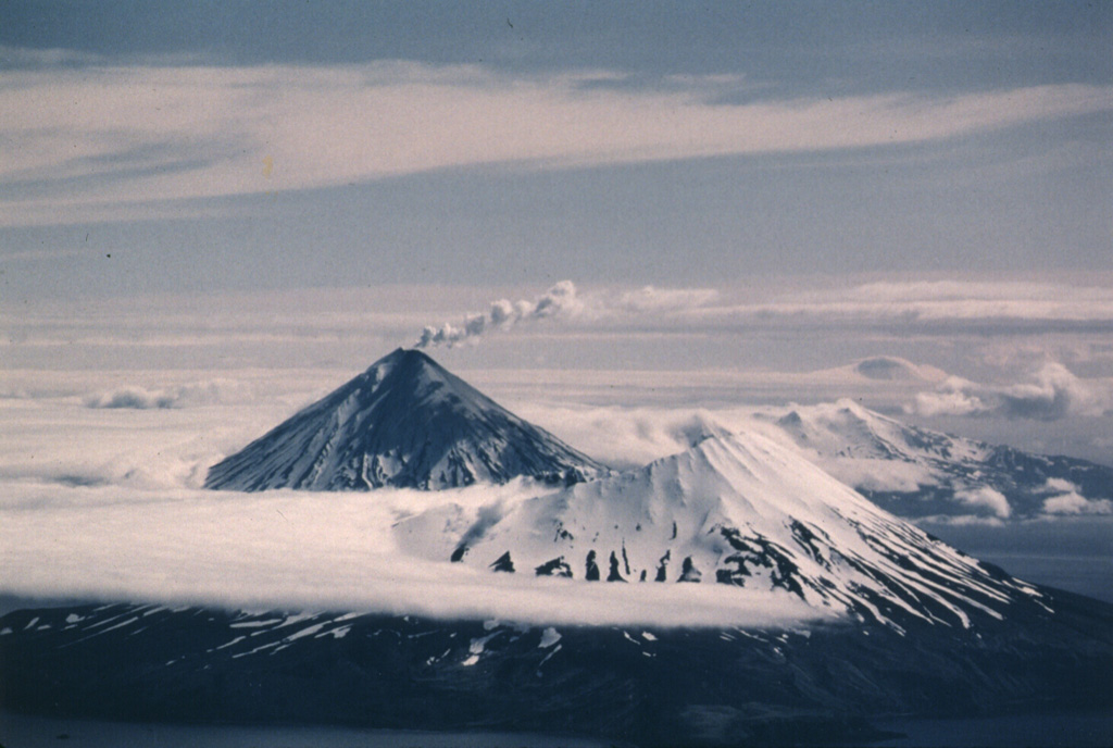 Herbert is a cone containing a 2-km-wide summit caldera that opens to NW. This 23 June 1987 view from the WSW shows a small plume originating from Cleveland in background, with darkened slopes from ashfall produced during an eruption that began on 19 June. Photo by Harold Wilson (Peninsula Airways), 1987 (courtesy of John Reeder, Alaska Div. Geology Geophysical Surveys).