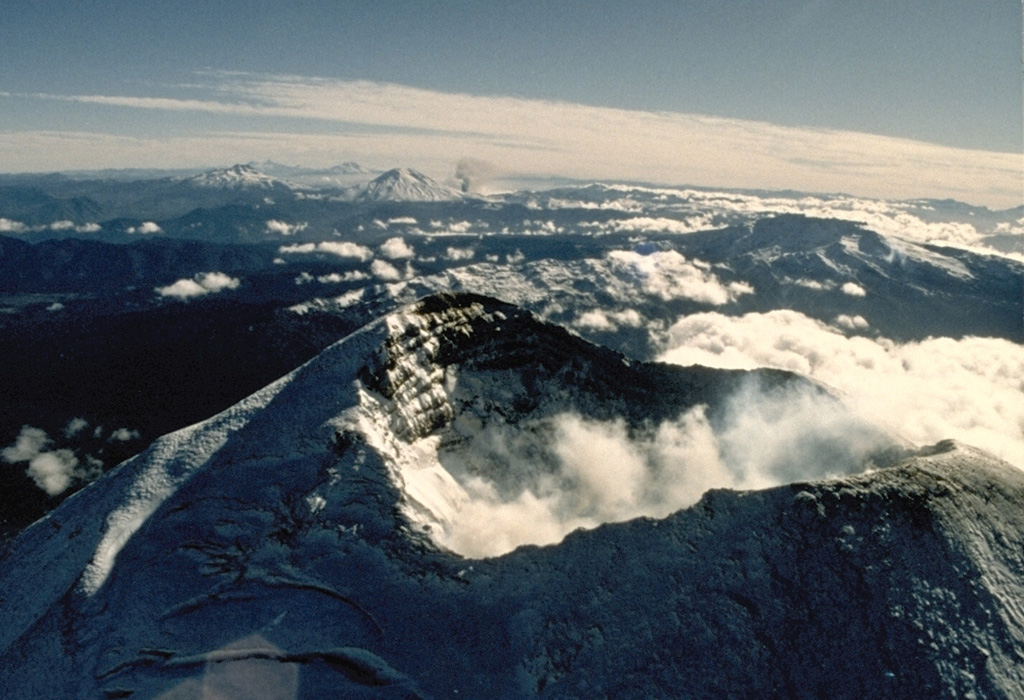 The summit crater of Llaima volcano appears in the foreground of this 1989 view looking north along the chain of Andean volcanoes.  An ash plume rises in the middle distance from a flank vent of Lonquimay volcano.  To its left is Tolguaca volcano, and Callaqui volcano lies farther to the north to the left of Lonquimay.  All these volcanoes except for Tolguaca have erupted in historical time; Llaima is one of Chile's most active volcanoes. Photo by Hugo Moreno, 1989 (University of Chile).