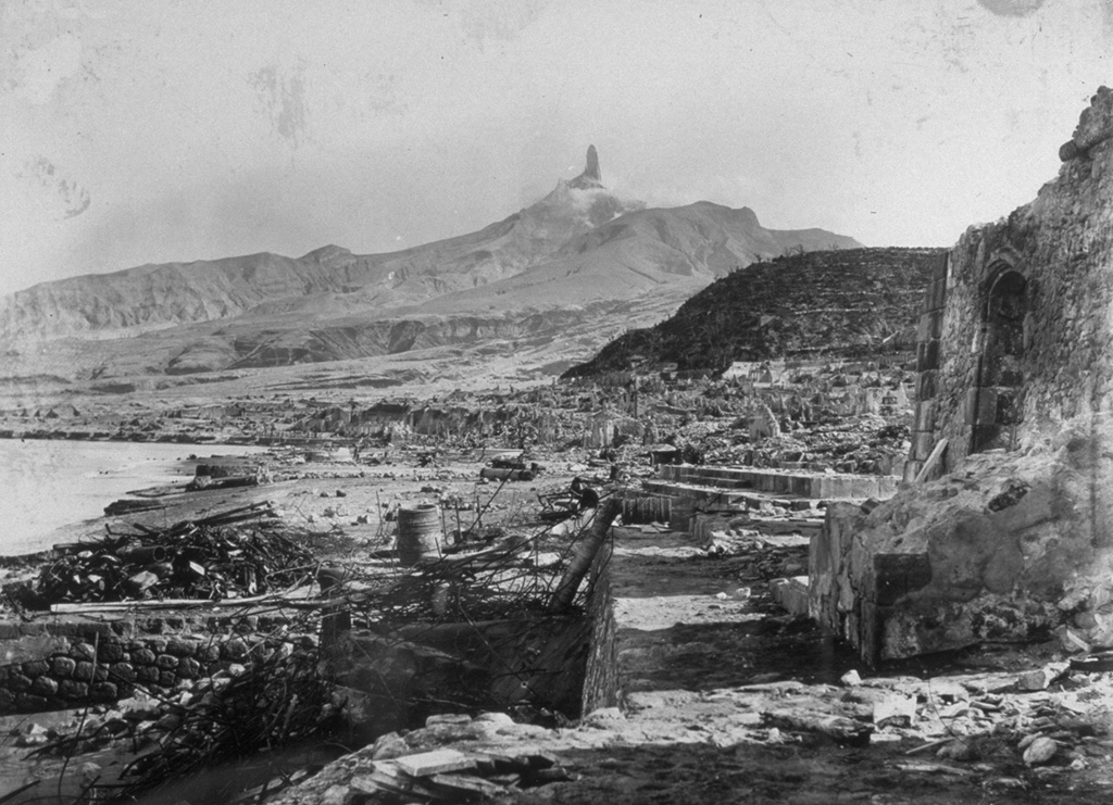 The devastated city of St. Pierre lies in ruins after a catastrophic eruption on 8 May 1902, in which pyroclastic flows and surges swept over the city, killing 28,000 people. The high-temperature pyroclastic surges devastated a 58 km2 area SW of the volcano and swept out to sea, capsizing all but two ships in the harbor.  This March 1903 photo from the south shows Mount Pelee towering over the remnants of the city, capped by a dramatic lava spine that grew above the summit lava dome.   Photo by A. Lacroix, 1903 (from the collection of Maurice and Katia Krafft).