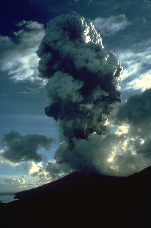 An eruption plume rises above the summit of Soufrière St. Vincent volcano on April 22, 1979.  A series of powerful explosive eruptions beginning on April 13 ejected the crater lake and removed the summit lava dome.  Pyroclastic flows occurred during several of the explosions and reached the west (lefthand) coast on April 14.  Following the cessation of explosive activity on April 25, a lava dome began growing in the new crater on May 3.  It eventually reached a height of 130 m and covered 840 m of the crater floor. Photo by Richard Fiske, 1979 (Smithsonian Institution).