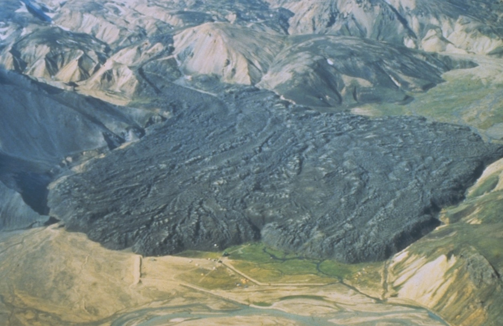 The viscous Laugahraun lava flow was emplaced in 1477 CE just inside the northern rim of Torfajökull caldera. This thick flow extends for about 2 km, and at its widest (left to right in this image), is about 1.3 km. Other small lava flows were produced at the same time as the Laugahraun lava flow: the Námshraun and Norðumámshraun lavas to the northeast just outside the caldera rim, and the Frostastaðahraun further north on the fissure system. The light-colored deposits surrounding the darker lava flow are rhyolitic volcanics. Photo by Richie Williams, 1981 (U.S. Geological Survey).