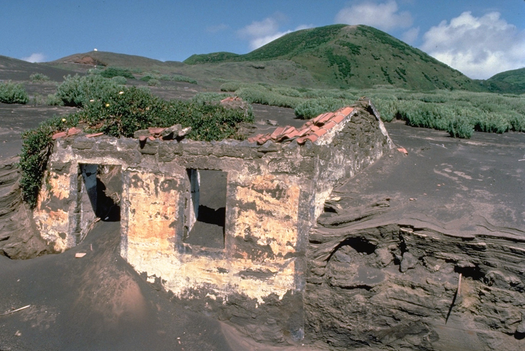 Accumulated thin layers of ashfall deposits reach nearly to the roofline at both gable ends of a house on the western tip of Fayal Island.  Repetitive, long-term ashfall during a 1957-58 eruption built an offshore cinder cone that added 1 km to the island's western peninsula. Copyrighted photo by Katia and Maurice Krafft, 1980.
