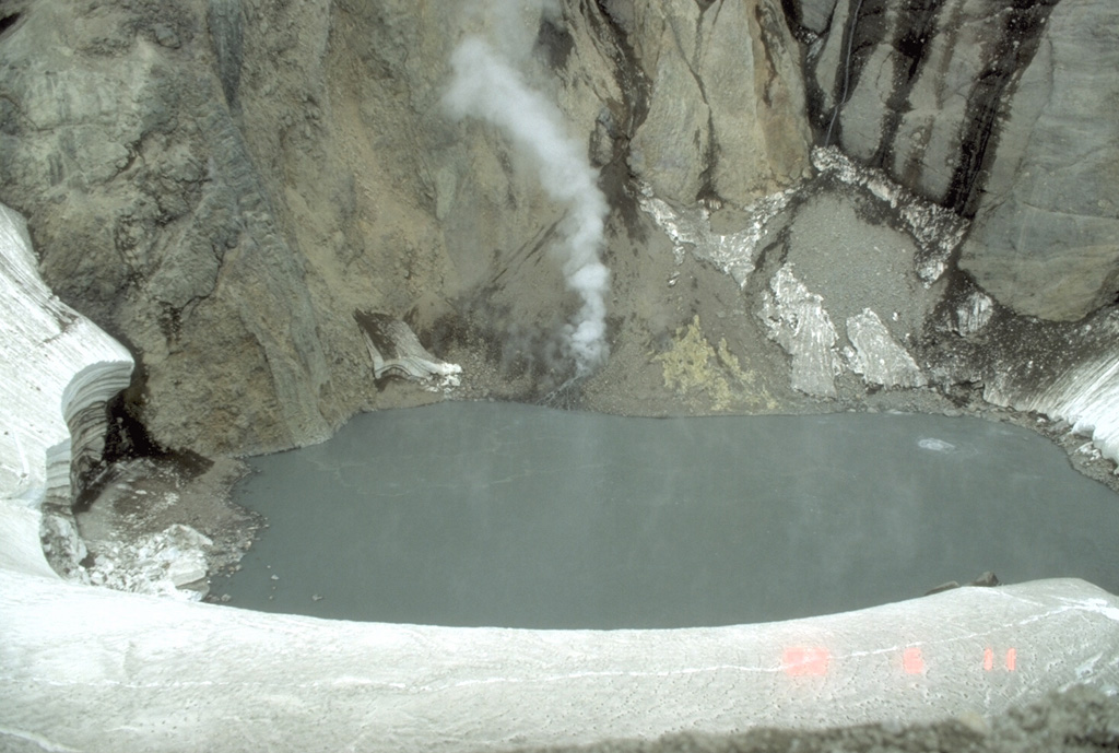 In early June 1992, this lake inside Crater Peak vent at Mount Spurr had a temperature of 49.7°C and a pH of 2.5. Note a circular upwelling zone about 5 m across to the upper right, and the plume rising from the talus pile on the far shore. This photo was taken on 11 June, a little more than two weeks prior to a major explosive eruption that ejected the crater lake.  Photo by Game McGimsey, 1992 (Alaska Volcano Observatory, U.S. Geological Survey).