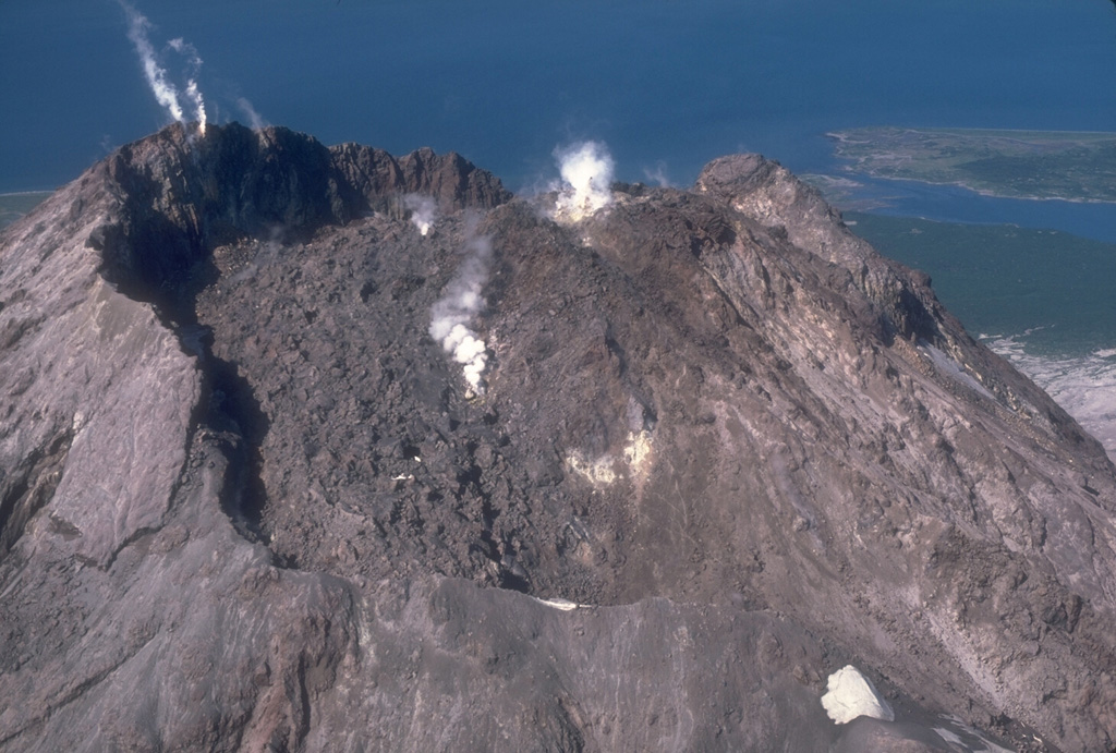 The 1976 lava dome, seen from the east in this 1982 photo of Augustine, formed within an arcuate crater rim that was produced by explosive removal of part of the 1964 lava dome during an earlier phase of the 1976 eruption. The small peak to the upper right is a remnant of the 1935 lava dome. Small plumes rise from the 1976 dome (center) and from fumaroles at the summit of the 1964 dome remnant (upper left). Photo by Chris Nye, 1982 (Alaska Division of Geological and Geophysical Surveys).