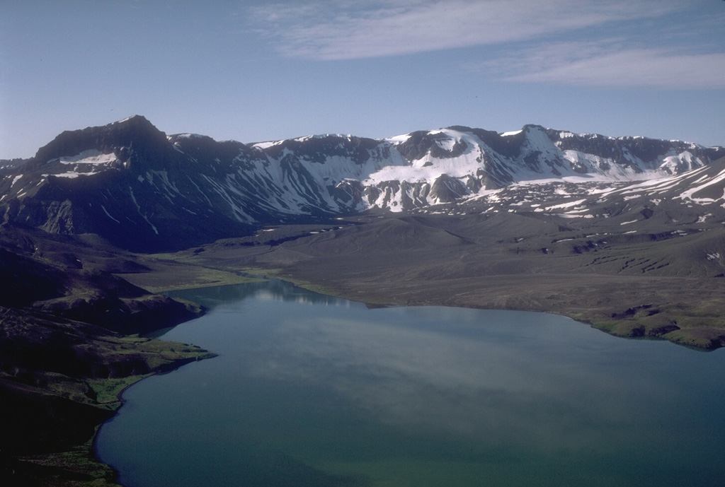 This view looks south across the caldera from the north rim of Aniakchak. The prominent dark peak on the skyline (left) is Black Nose, a remnant of pre-caldera volcaniclastics. A pumice-covered glacier and associated moraine is in the distance against the caldera wall. Surprise Lake once covered a much larger part of the caldera floor before catastrophically draining through a notch in the east caldera rim. Photo by Tom Miller, 1985 (Alaska Volcano Observatory, U.S. Geological Survey).