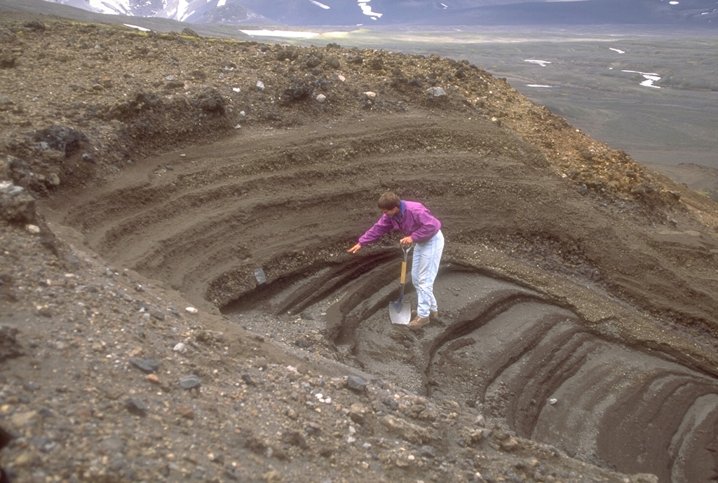 A geologist examines pyroclastic deposits (above hand) from a violent eruption of Half Cone less than 500 years ago. The Half Cone layers overlie dark gray phreatomagmatic deposits from Surprise tuff cone in Aniakchak caldera. The Half Cone eruption produced about 1 km3 of tephra and resulted in truncation of the SE side of the cone. Photo by Game McGimsey (Alaska Volcano Observatory, U.S. Geological Survey).