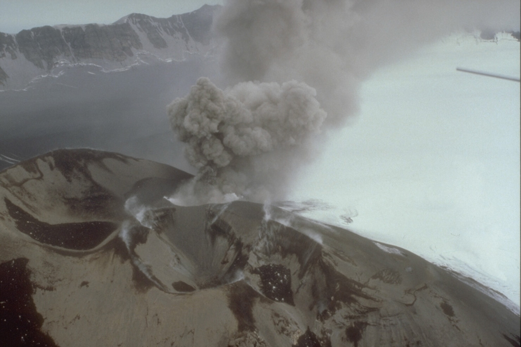 In this 3 August 1993 photo a small ash plume rises from a scoria cone within the Veniaminof caldera. Intermittent explosive eruptions began on 30 July and continued for more than a year. The eruptions also formed a new cone SE of the main western cone. As during the 1983-84 eruption, lava flows erupted onto the ice-covered caldera floor. Ash darkens the left side of the caldera floor in this view, with the caldera wall in the background. Photo by D. Sellers, 1993 (Alaska Department of Fish and Game).
