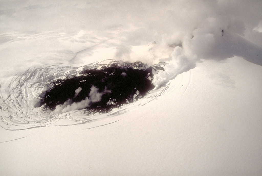 On 9 May 1994, a fresh black lava flow is erupted onto the glacier-covered caldera floor of Veniaminof volcano on the Alaska Peninsula. The source of the lava flow is a cinder cone that is obscured by steam to the upper right. Concentric fractures on the glacier surface are created as the lava flow melts through the glacial ice.  Photo by Chris Nye, 1994 (Alaska Division of Geological and Geophysical Surveys).